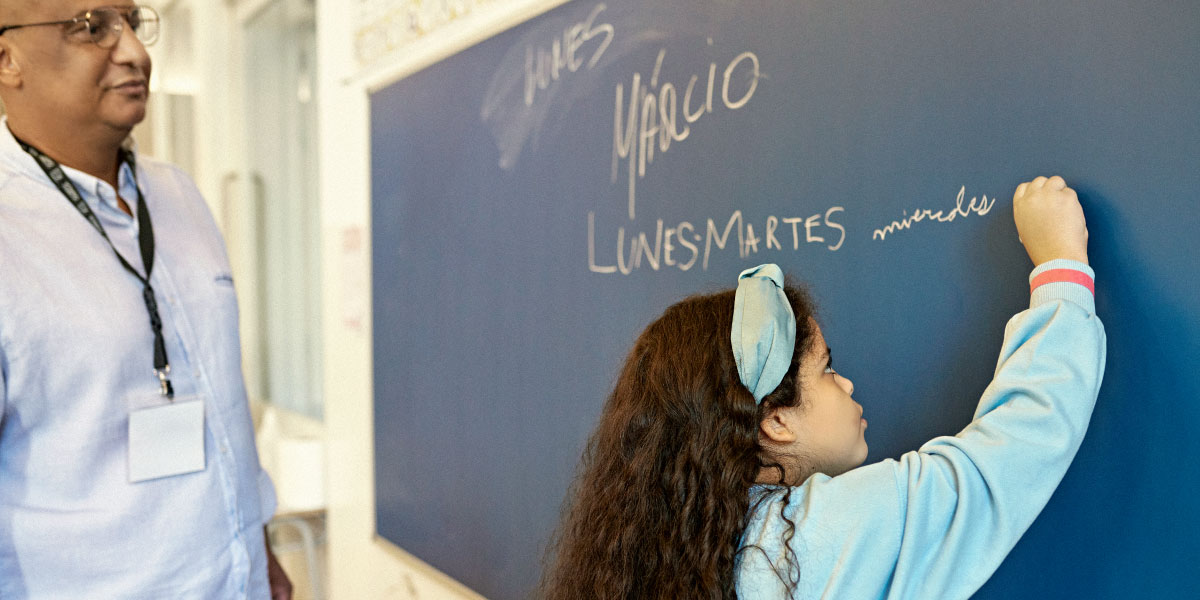 A student writing in spanish on a chalkboard while their teacher watches