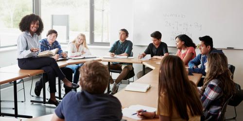 A teacher speaking to a small group of students sitting with their desks in a circle